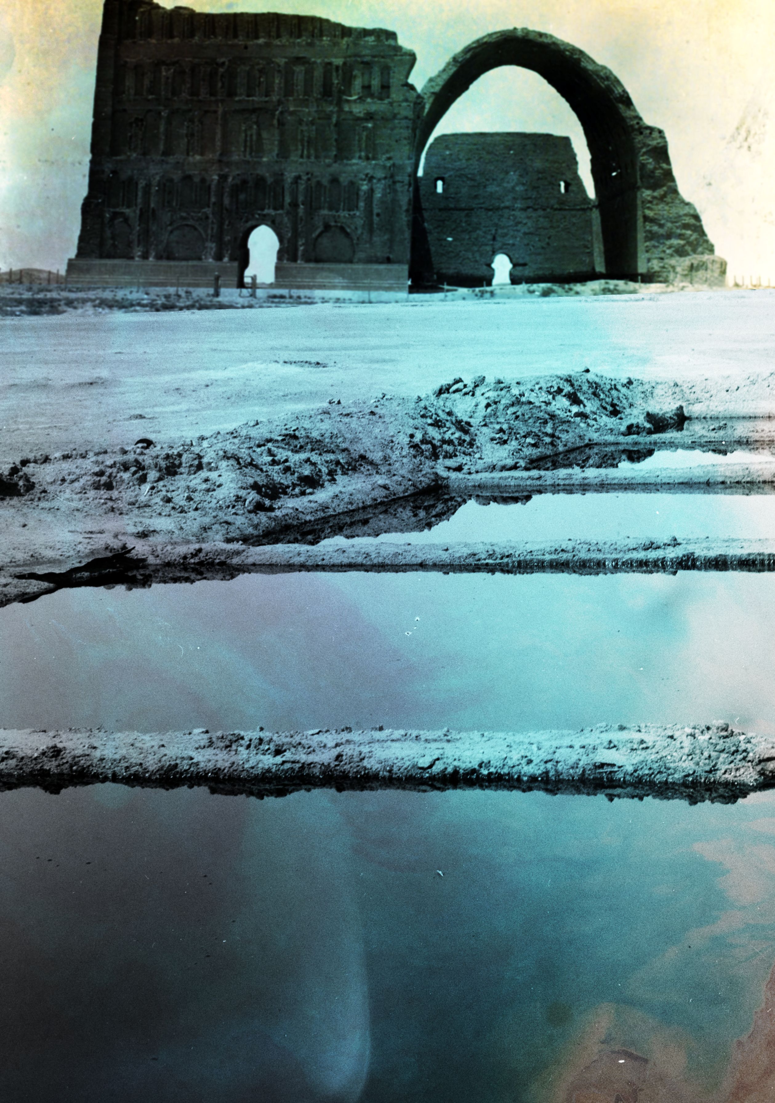 foreground, rectangular pools with mirror-like cyan liquid with indigo and ochre marbling receding towards the top right, giving way to light blue sands. background, a ruined building consisting of a monumental face and an ovoid arch, coloured green and dark turquoise, stands against a pastel peach sky. the entire image has the grain of an old photograph